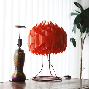En retroinspirerad orange bordslampa med en unik geometrisk lampskärm och en färgglad vaslampa i närheten, placerad på en träyta med en modern bakgrund av vita persienner och växter.